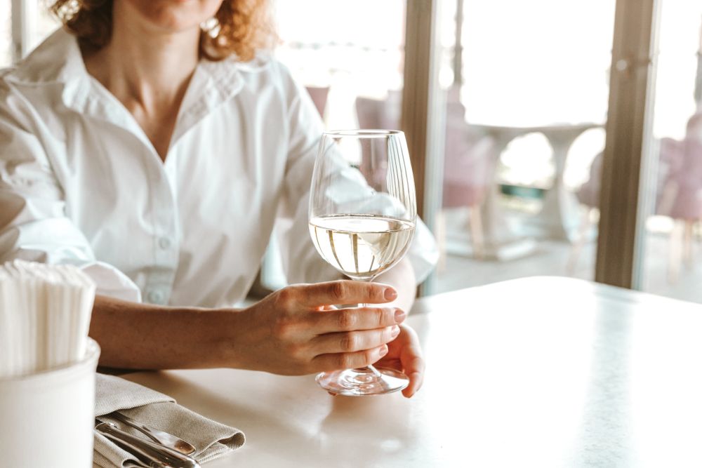 Gošća restorana drži čašu belog vina za stolom pored prozora.